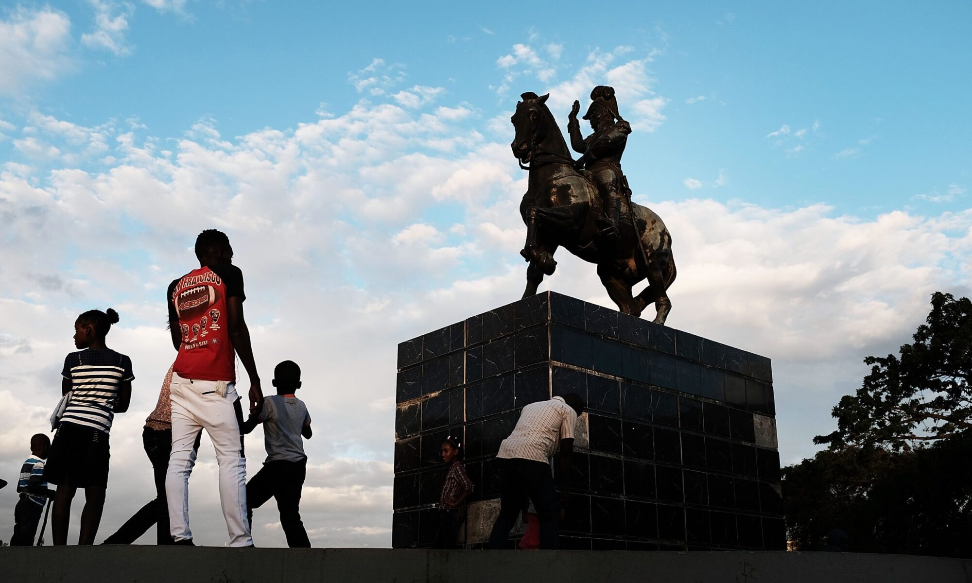 People stand under a statue of Jean-Jacques Dessalines, a leader of the Haitian Revolution and the first ruler of an independent Haiti, on February 10, 2018, in Port-au-Prince, Haiti. Photo by Spencer Platt/Getty Images.